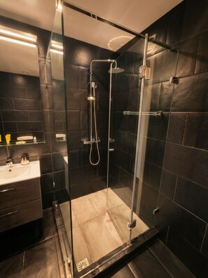 A modern bathroom with a glass-enclosed shower, dark tiled walls, and a visible sink area. - Elite Ermou Suites (Athens)