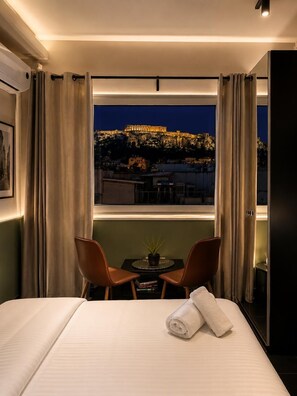 A hotel room with a bed, two chairs, a small table, and a view of a cityscape at night. - Elite Ermou Suites (Athens)