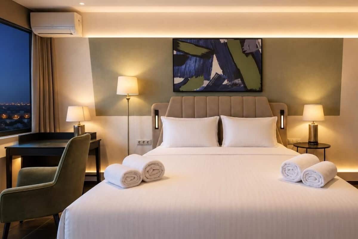 A hotel room with a bed, two bedside lamps, a desk with a chair, and a view of the city at night.