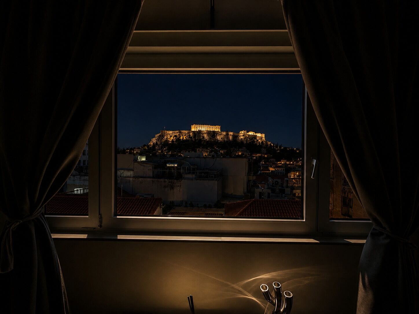 A view of a historic building illuminated at night, seen through a window with curtains.