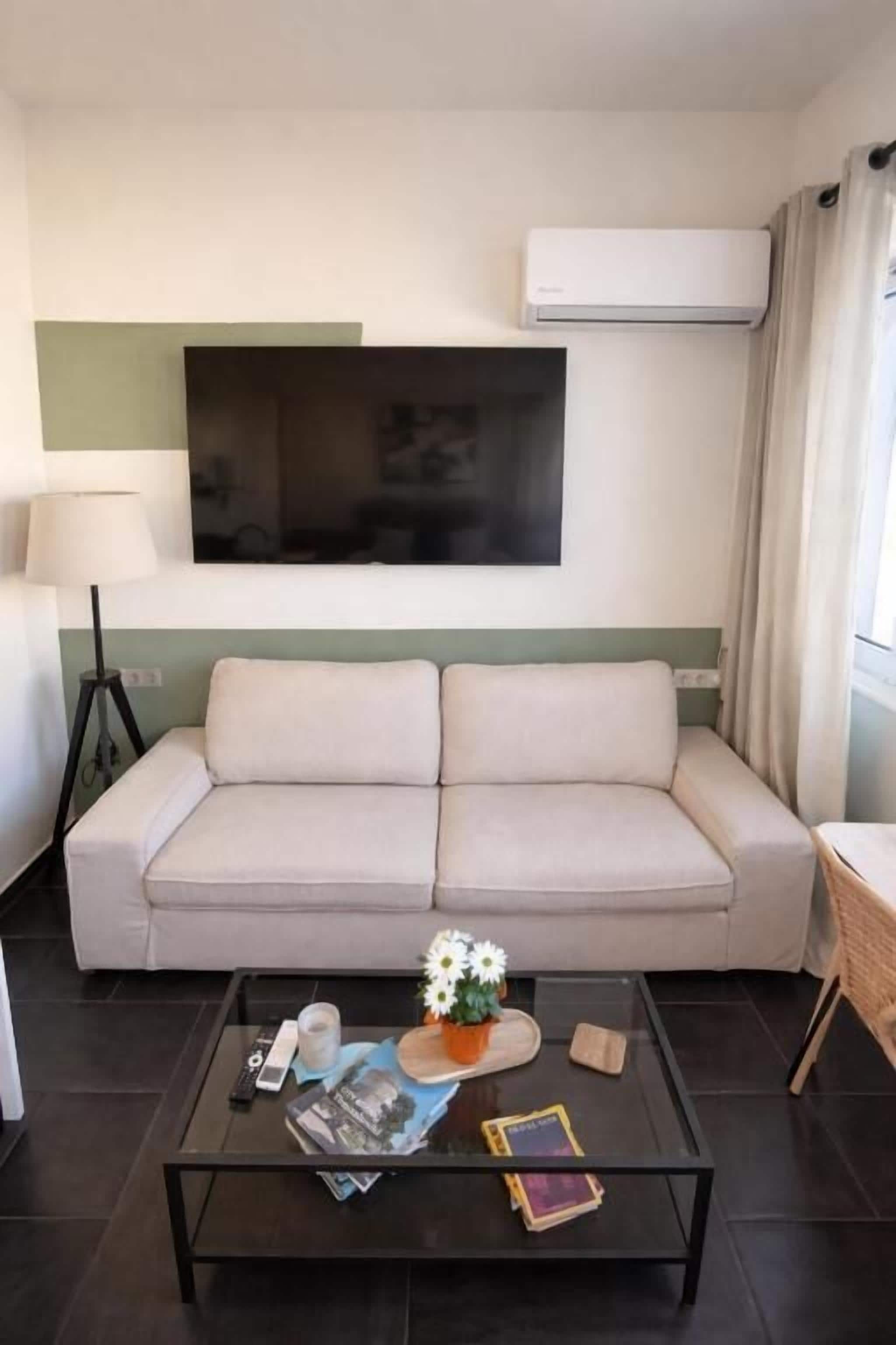 Una sala de estar con un sofá, una mesa de centro con libros y un florero, una lámpara y un televisor de pantalla plana montado en la pared.