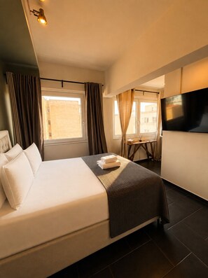 A hotel room with a bed, curtains, a TV, and a view of the outside. - Elite Ermou Suites (Athens)