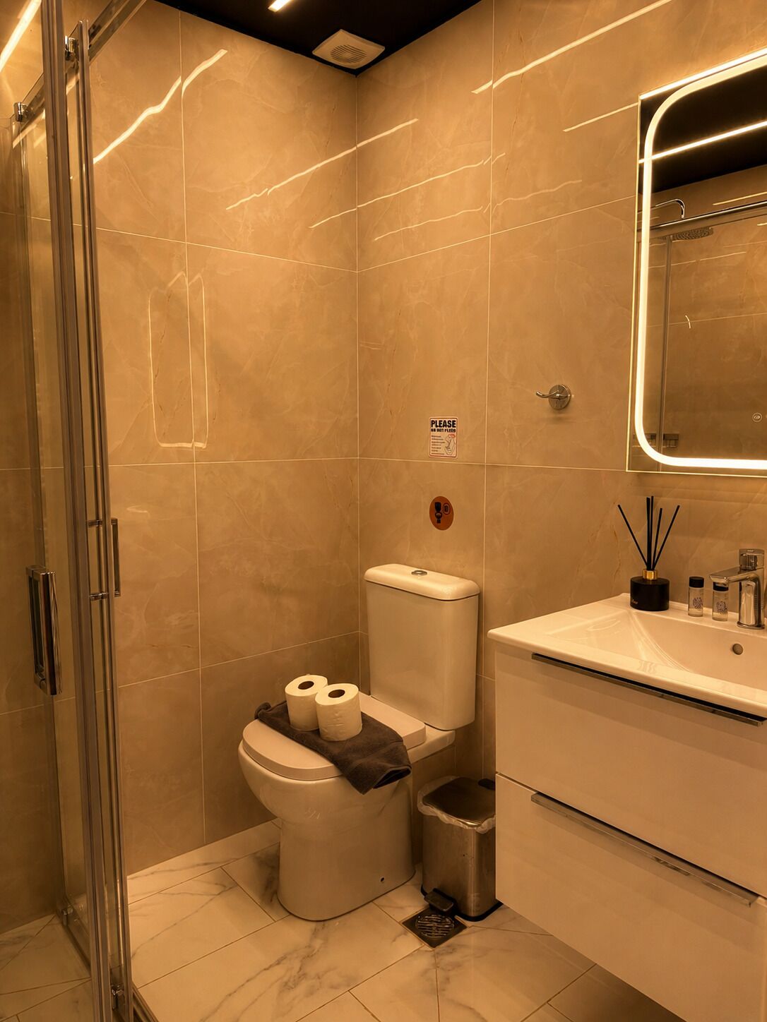 Et moderne badeværelse med en glasbrusekabine, toilet med toiletpapirsholder, skraldespand og et håndvask med spejl over.