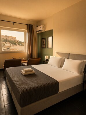 A hotel room with a large bed, a desk, a chair, a window with a view of a cityscape, and an air conditioning unit. - Elite Ermou Suites (Athens)