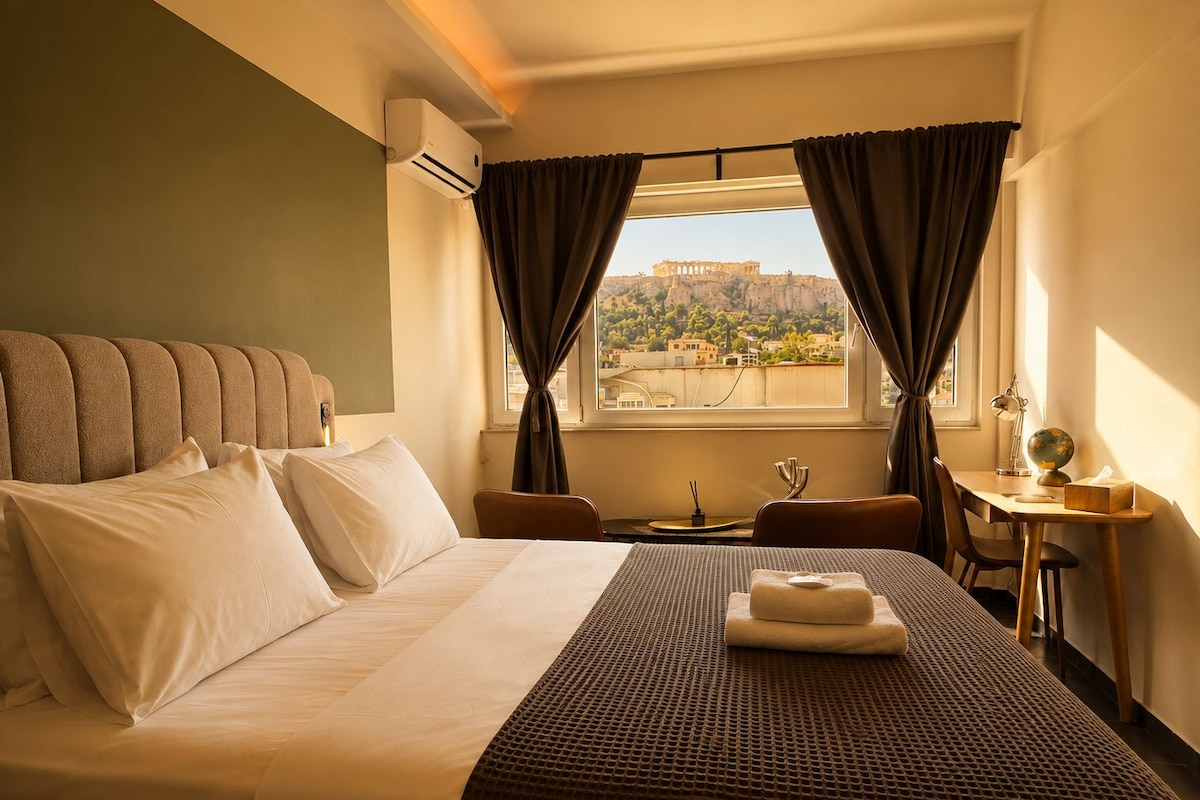 Quarto de hotel com cama, escrivaninha, cadeira, janela com cortinas e vista para um edifício histórico.