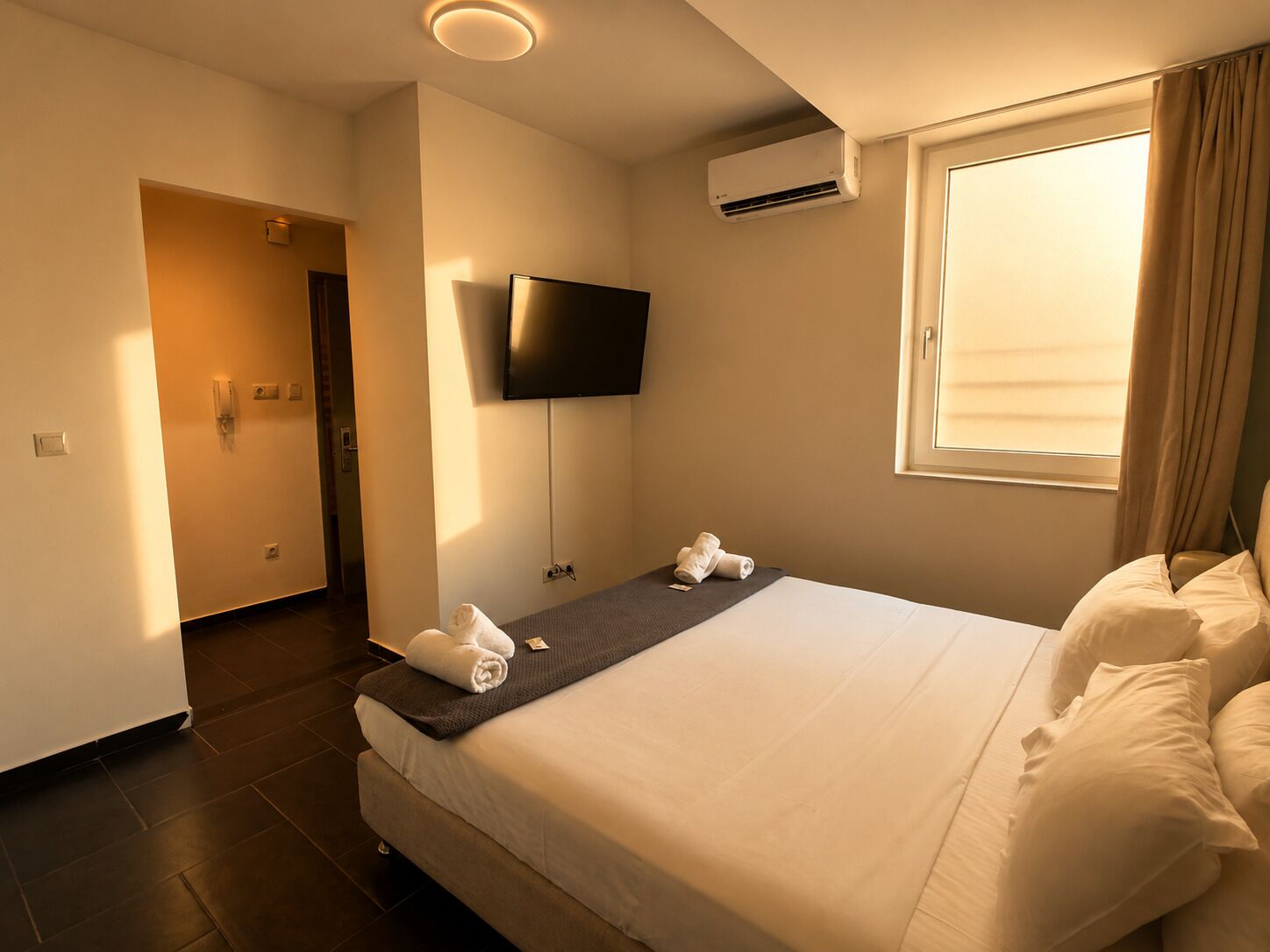 A hotel room with a bed, a flat-screen TV mounted on the wall, an air conditioning unit, and a window with curtains.