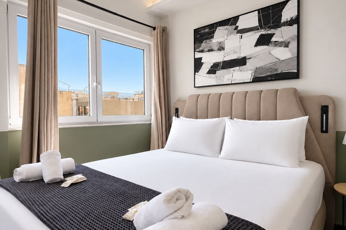 A hotel room with a bed, a window with curtains, a painting on the wall, and a view of buildings outside.