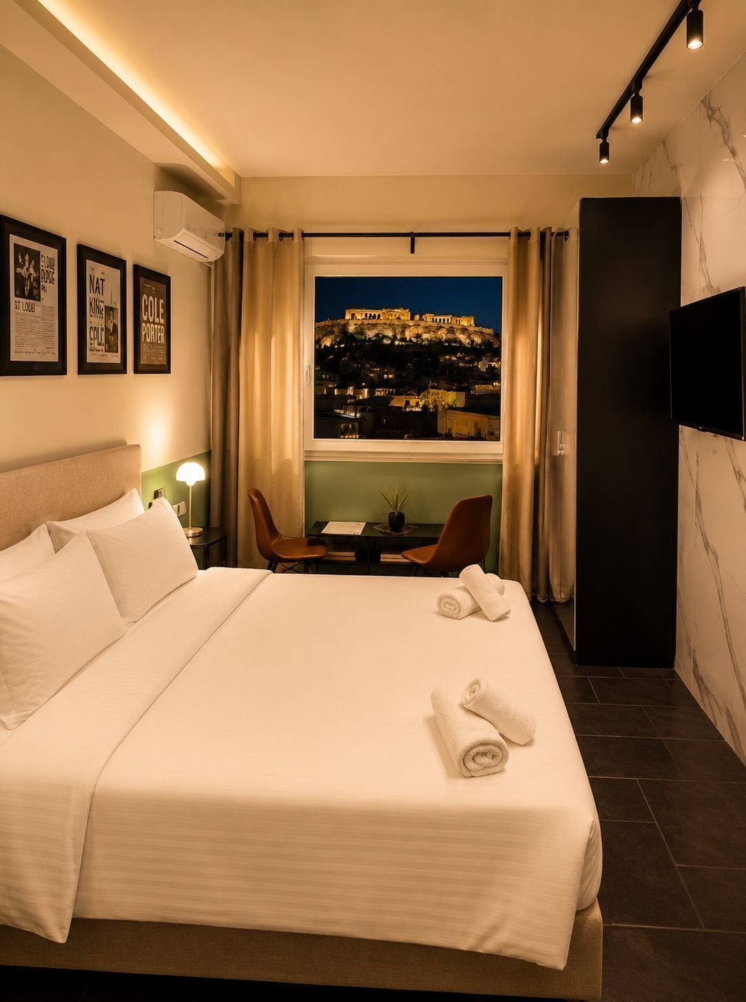 A hotel room with a bed, a desk with chairs, a TV, and a view of a cityscape at night.