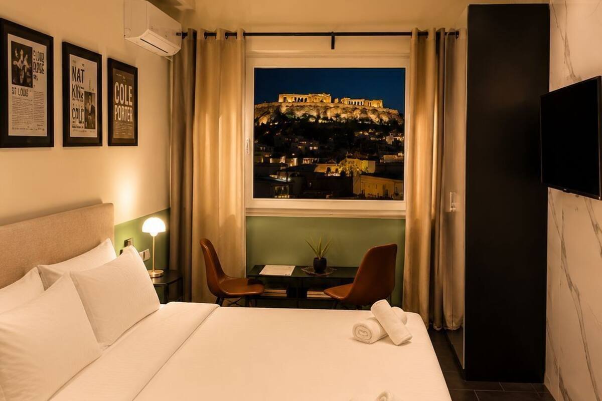 A hotel room with a bed, a desk with chairs, a TV, and a view of a cityscape at night.