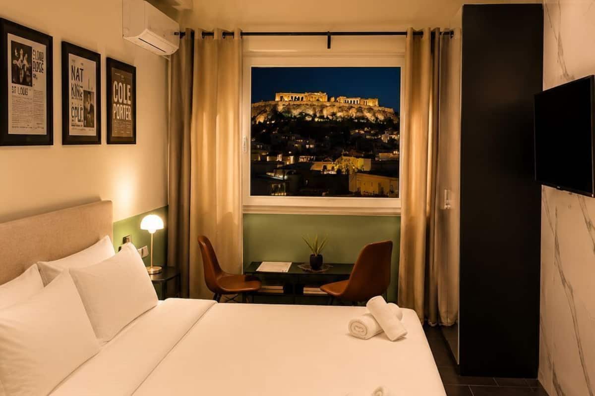 A hotel room with a bed, a desk with chairs, a TV, and a view of a cityscape at night.