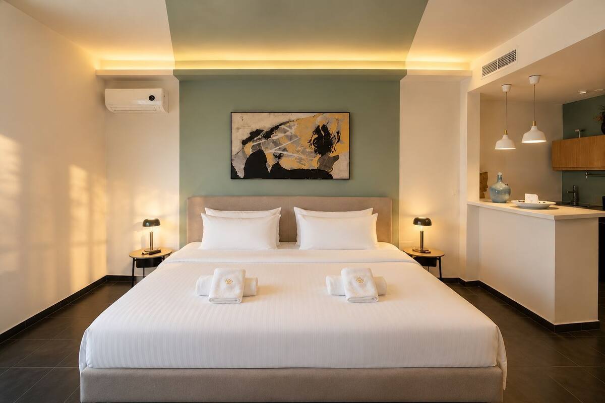 A hotel room with a bed, two bedside tables, a painting on the wall, a kitchenette, and an air conditioning unit.