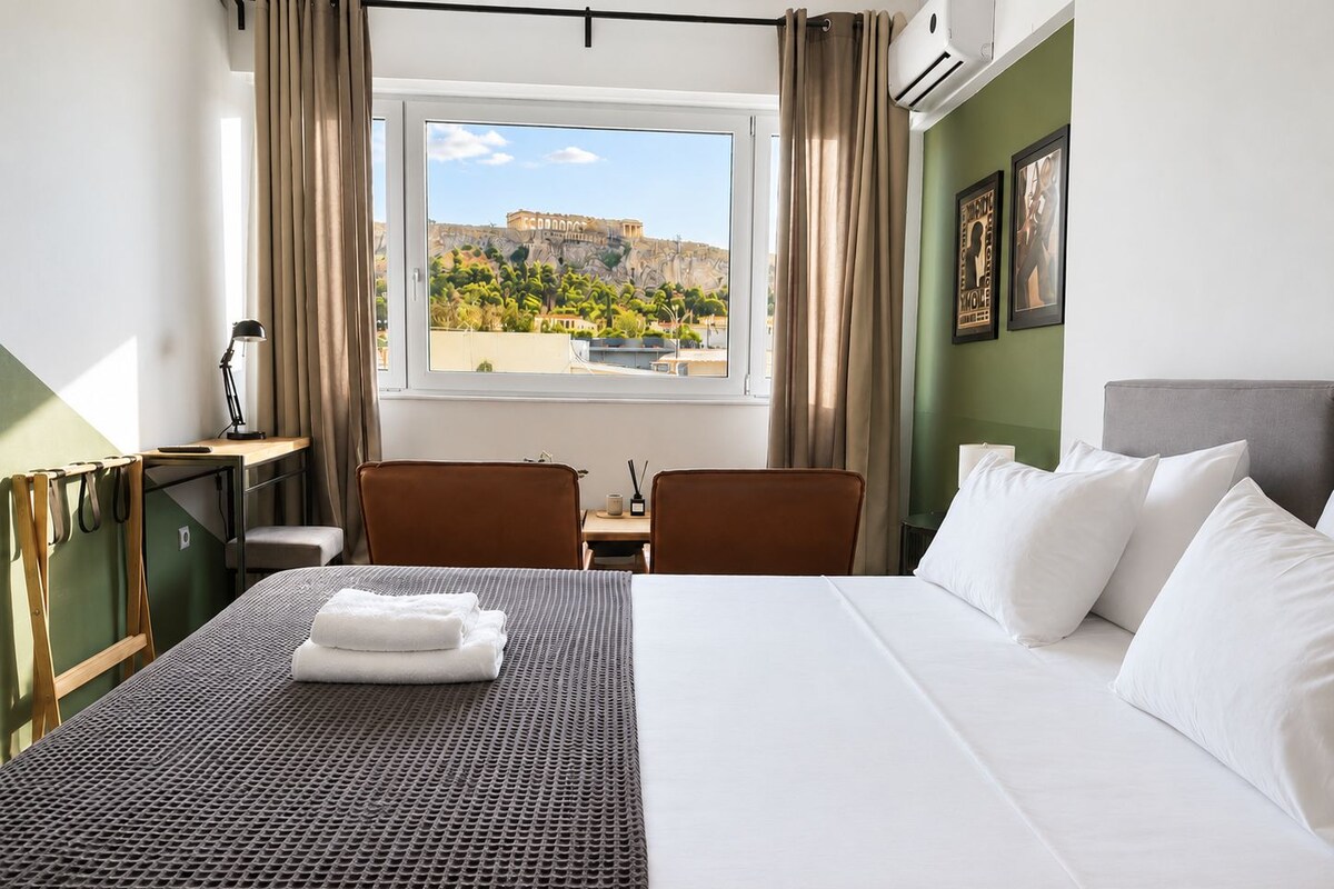 A hotel room with a bed, two chairs, a desk, a window with a view of a historic building, and a painting on the wall.