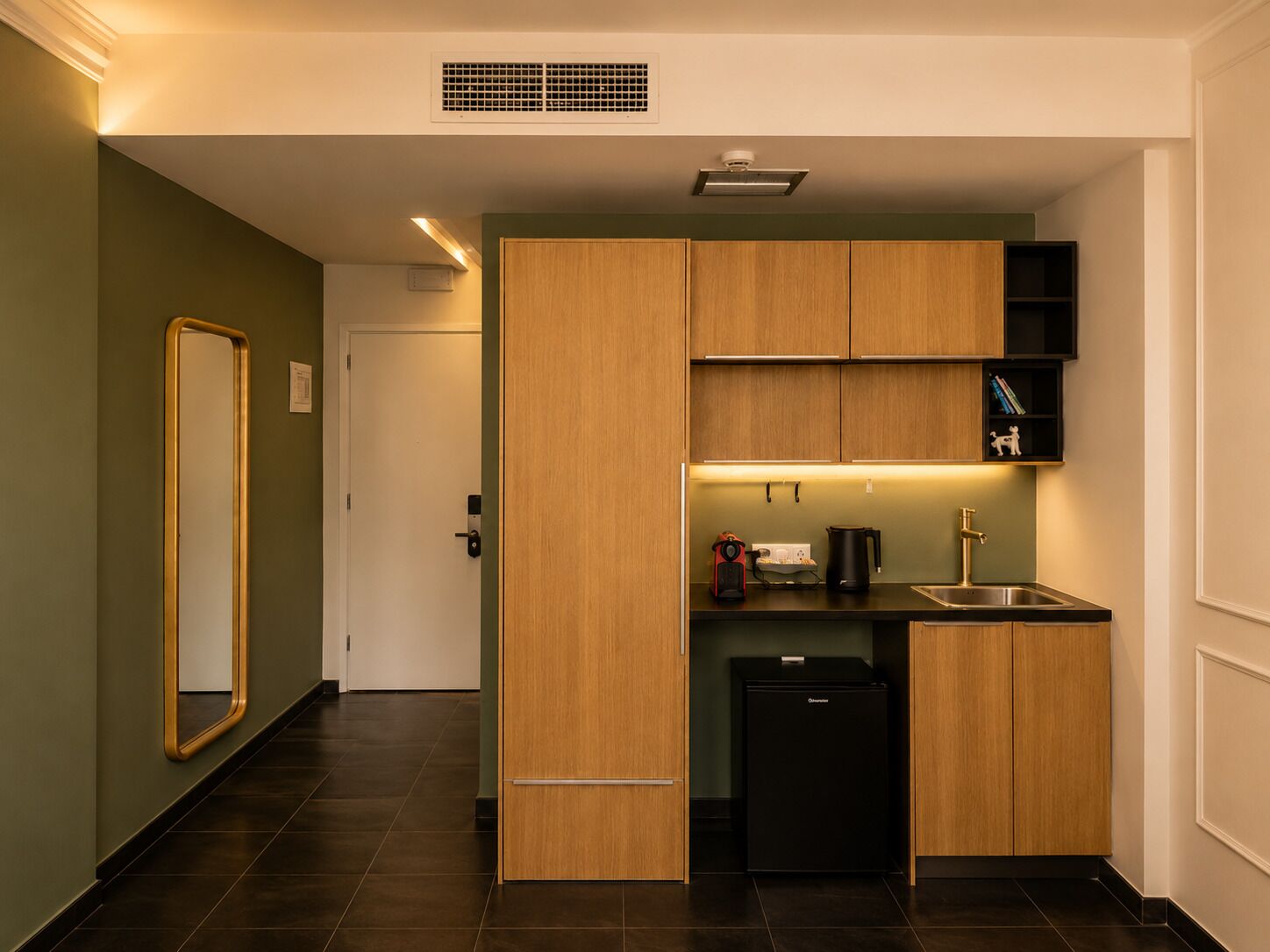 Cocina compacta con armarios de madera, un refrigerador negro, un lavamanos y una encimera con hervidor y cafetera.