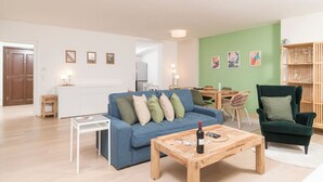 A living room with a blue sofa, a wooden coffee table, and a green armchair. There is a dining area with a table and chairs, and a kitchen in the background. - Premium Business Apartment (Vienna)