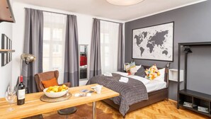 Ein modernes Hotelzimmer mit Bett, Esstisch mit Frühstück, Stuhl, einer Weltkarte an der Wand und einem großen Fenster mit Vorhängen.