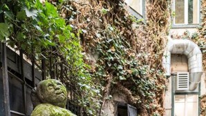 Eine Steinstatue einer Figur in einer Gartenanlage mit einer Steinmauer und blühenden Pflanzen.