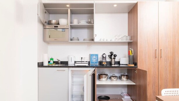 A compact kitchen with white cabinets, a built-in oven, and a small refrigerator. There are shelves above the counter holding dishes and glasses. - Apartment Favoriten top 1 (Vienna)