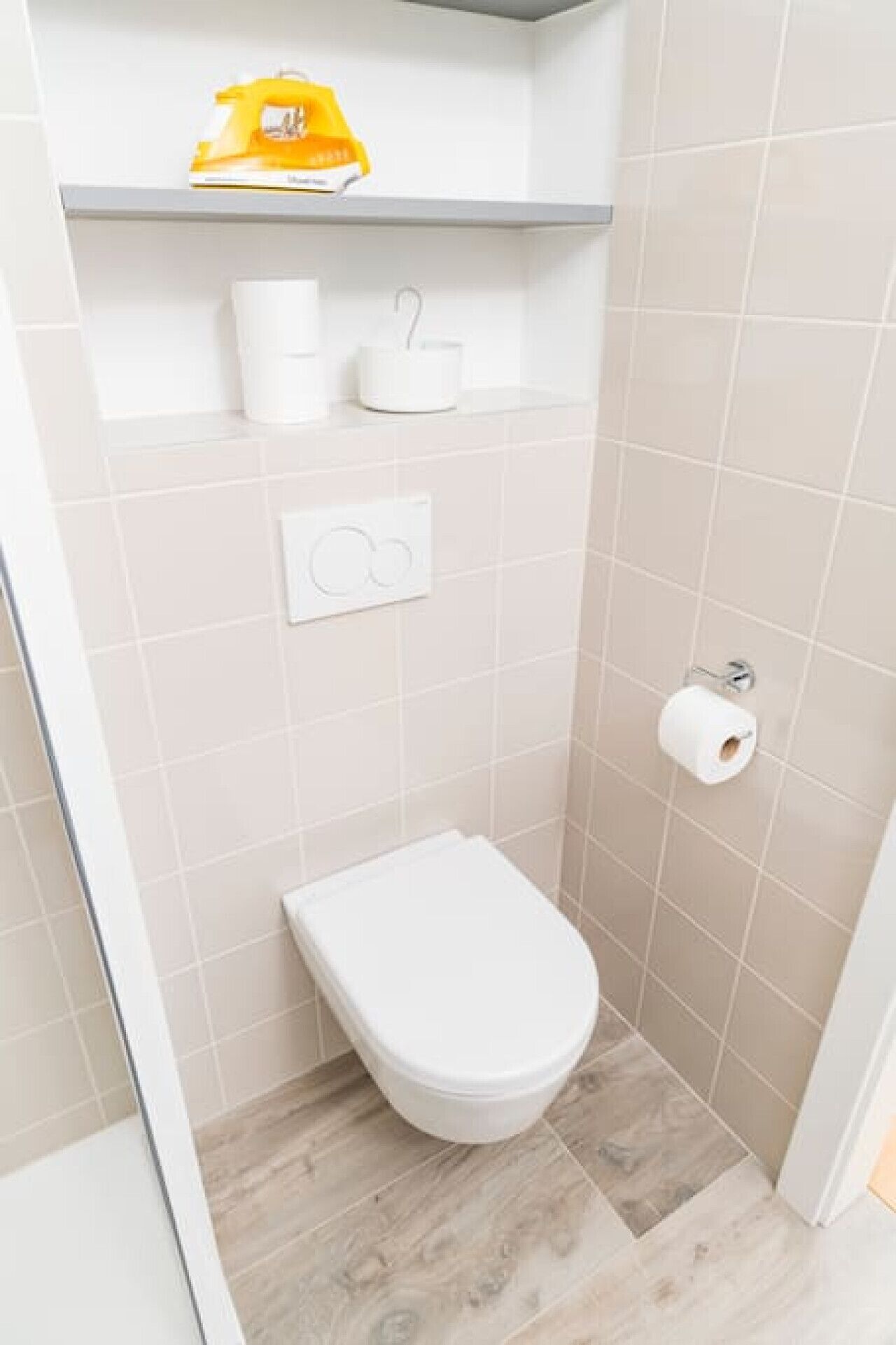 Een kleine badkamer met een toilet, een plankje met een geel voorwerp, en een rol toiletpapier.