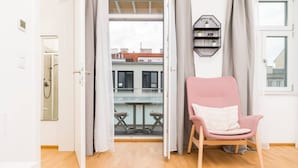 Een open deur die leidt naar een balkon met een tafel en stoelen, een roze stoel binnen en een zwarte plank met planten.