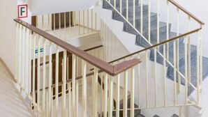 Una escalera en espiral con barandillas blancas y pasamanos de madera, que lleva a un rellano donde se encuentra un extintor montado en la pared.