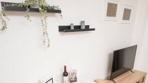 Un comedor con una mesa blanca, dos sillas blancas, un cuenco de fruta, una botella de vino y un televisor sobre un estante de madera.