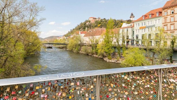 Une rivière traversée par un pont, bordée de bâtiments et d’une rambarde métallique ornée de cadenas.