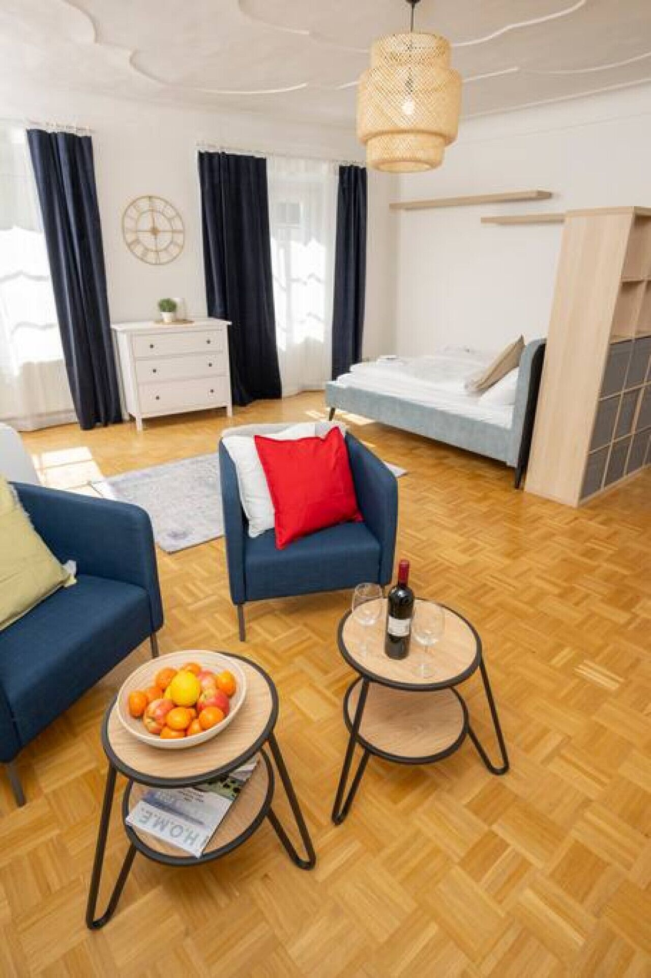 A modern living room with a sofa, armchair, and coffee table. There is a bed in the background and a window with curtains.