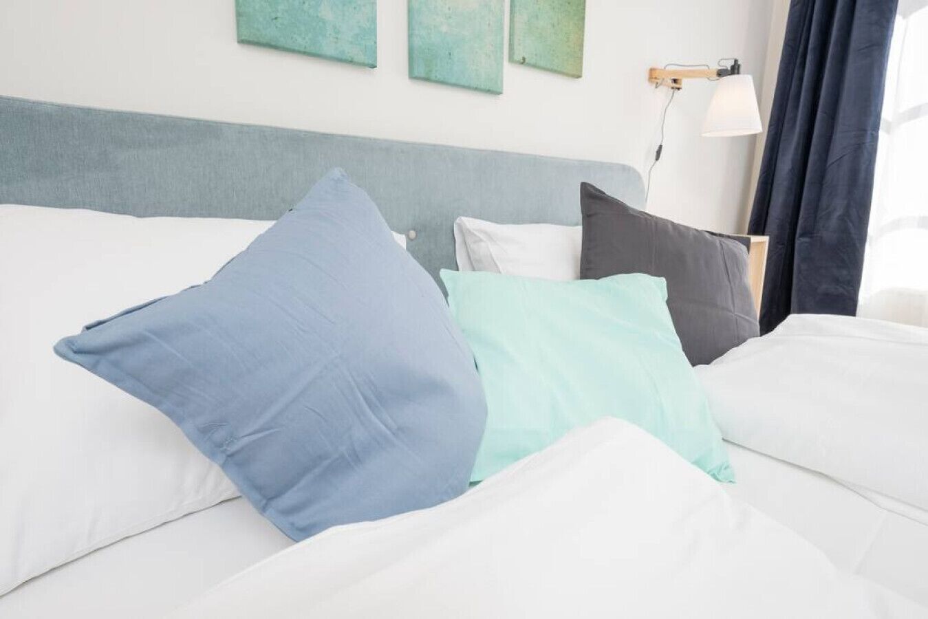 Ein Bett mit weißer Bettwäsche und drei Kissen in Blau, Hellblau und Grau.