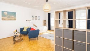 Un salon moderne avec un plancher en bois, un canapé bleu, une petite table sur laquelle se trouve une bouteille, et une grande fenêtre avec des rideaux.