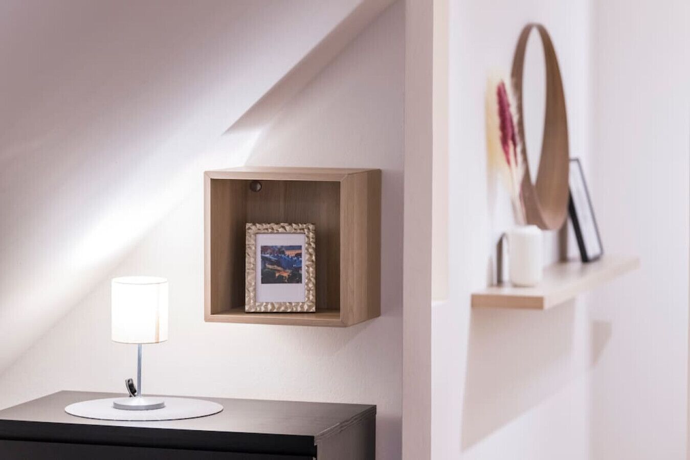 Ein modernes Zimmer mit einem hölzernen Regal, das ein gerahmtes Bild, eine weiße Lampe auf einem schwarzen Tisch und einen Spiegel auf einer unteren Ablage zeigt.