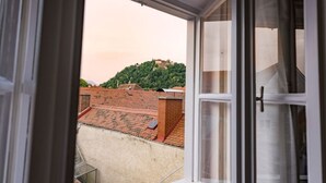 Vista da janela sobre um terraço com um edifício de tijolos e uma colina com vegetação.