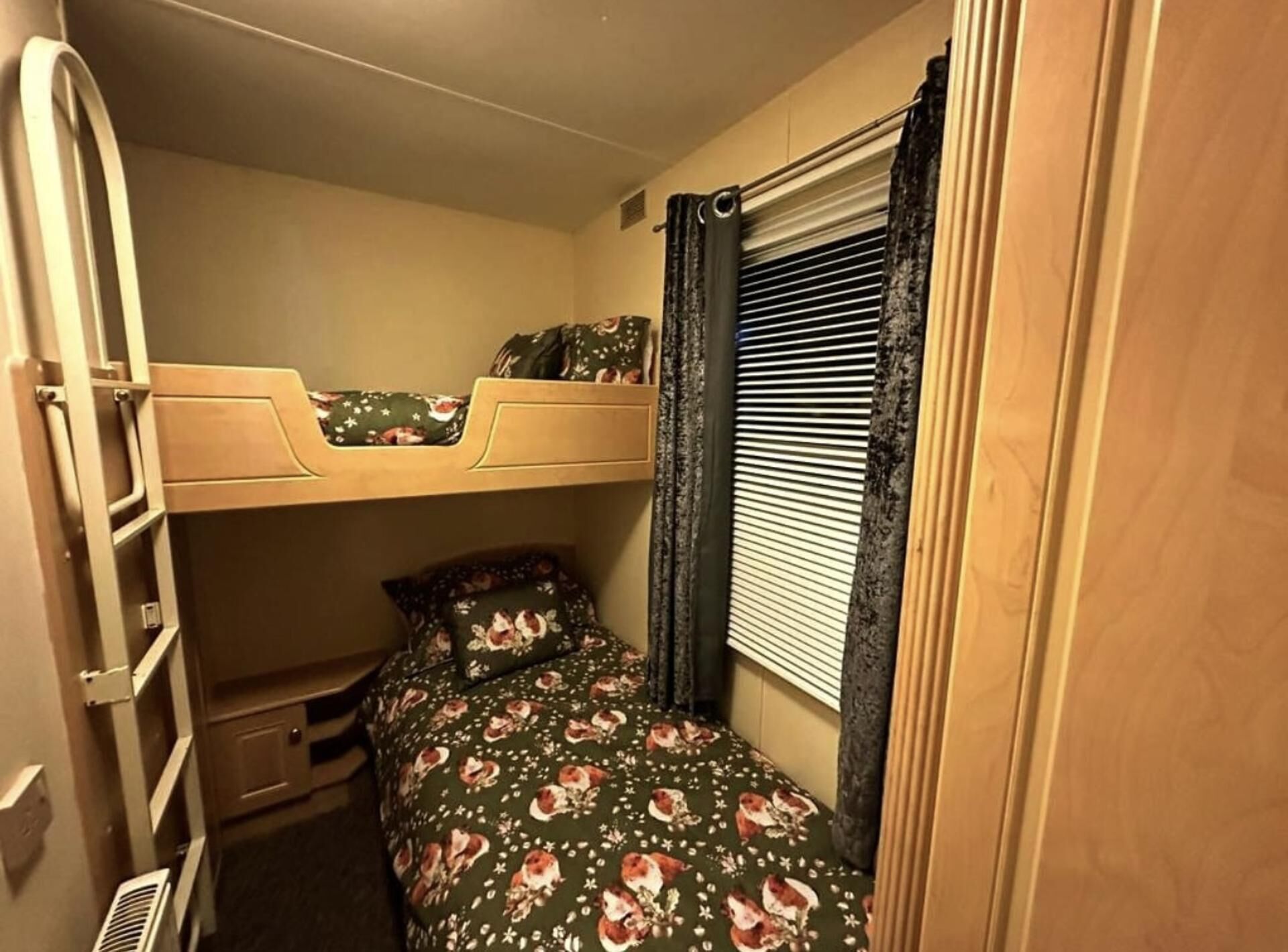 Ein Stockbett mit einer unteren Schlafgelegenheit und einem kleineren Bett darunter. Ein Fenster mit Jalousien und einem Vorhang ist vorhanden.