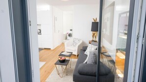 A modern living room with a grey sofa, a small coffee table, and a white armchair. There is a washing machine in the background, and a glass door leading to another room. - Ottakring Balcony Suite (Vienna)