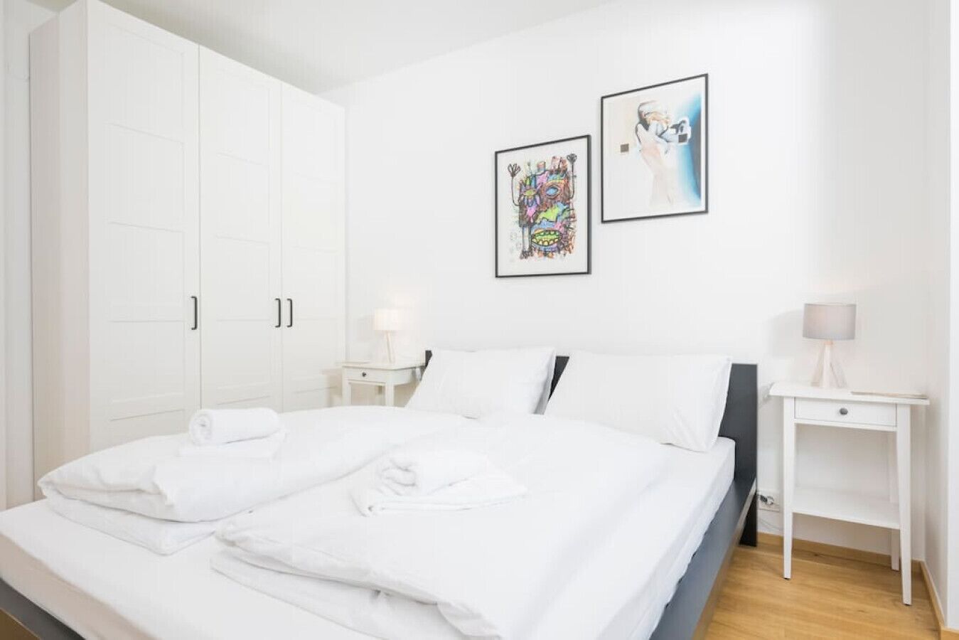 Une chambre d’hôtel avec un grand lit, des murs blancs, deux œuvres d’art encadrées et une petite table de chevet avec une lampe.