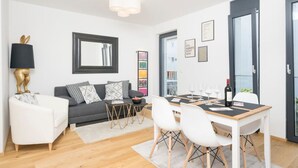 A modern living room with a grey sofa, white armchairs, a dining table with chairs, and a large window with a view. - Ottakring Balcony Suite (Vienna)