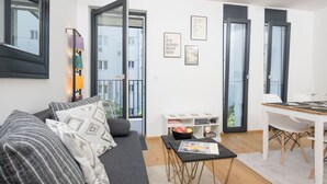 A modern living room with a sofa, coffee table, and a dining area with a table and chairs. There are open doors leading to a balcony, and a bookshelf with books and decorative items. - Ottakring Balcony Suite (Vienna)