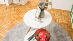 A room with a wooden herringbone floor, a white round table with a vase and a bowl of fruit, a grey rug, and a bed in the background. - Zum Grazer Uhrturm in Graz Austria (Graz)