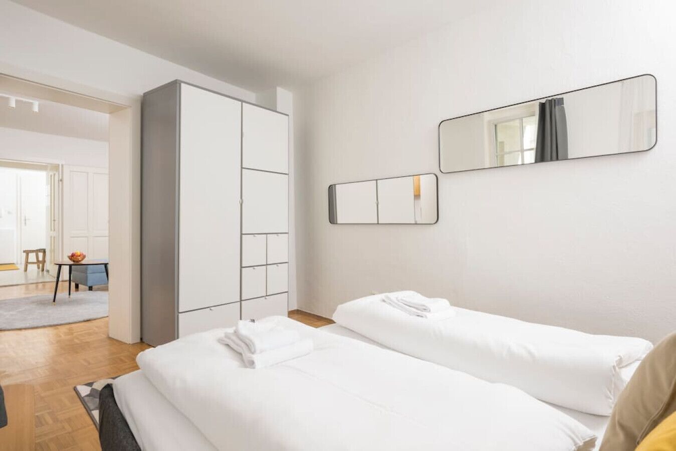 Une chambre d’hôtel avec deux lits, une armoire, une petite table sur laquelle se trouve un saladier rempli de fruits, et un miroir au mur.