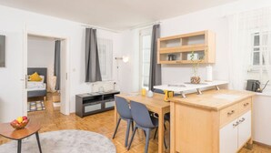 Ein modernes, helles Zimmer mit einem Holztisch, Stühlen, einem kleinen Tisch mit Obst und einem Bett mit einem gelben Kissen.