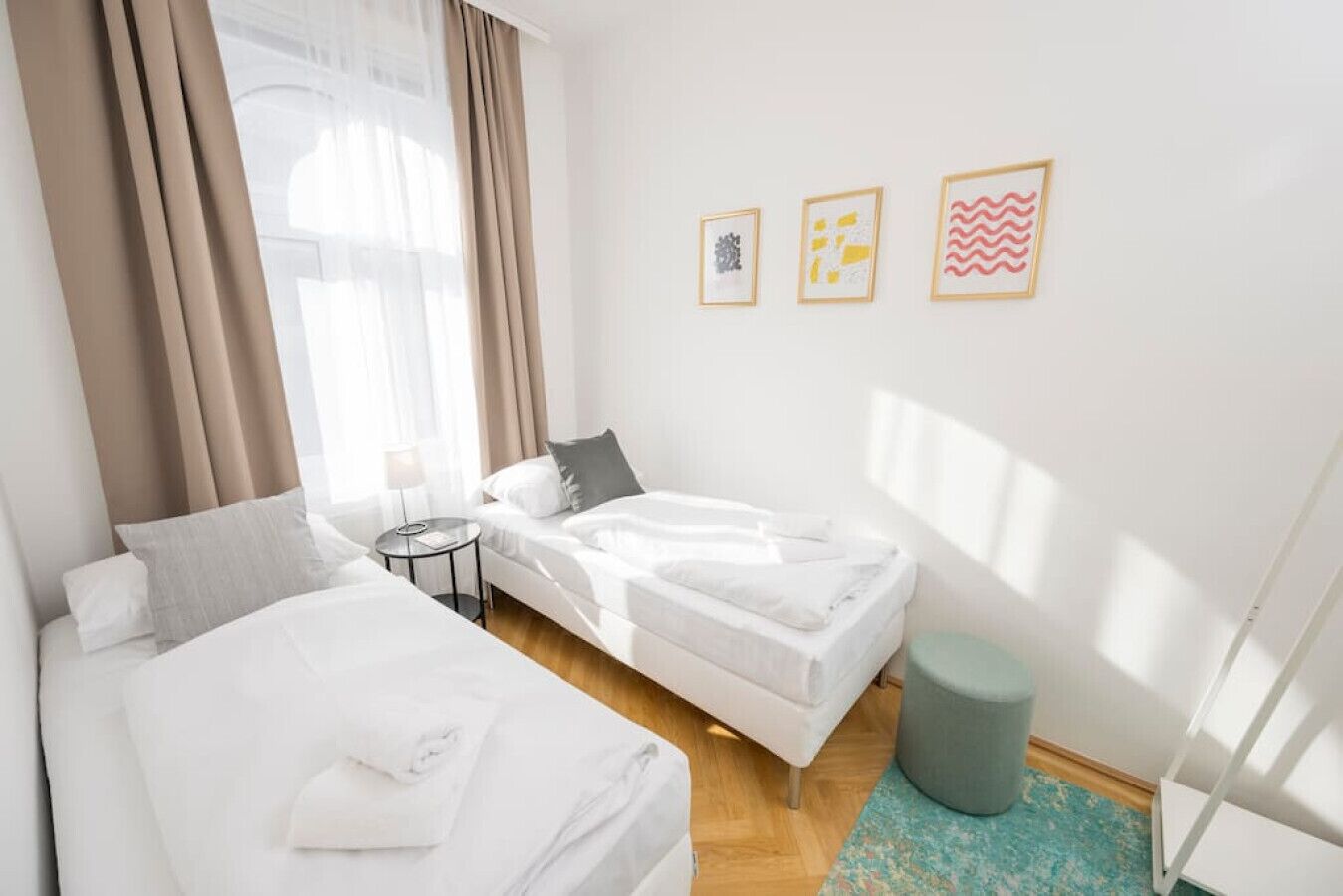 Een hotelkamer met twee bedden, een kleine ronde tafel, een grijze poef en drie ingelijste kunstwerken aan de muur.