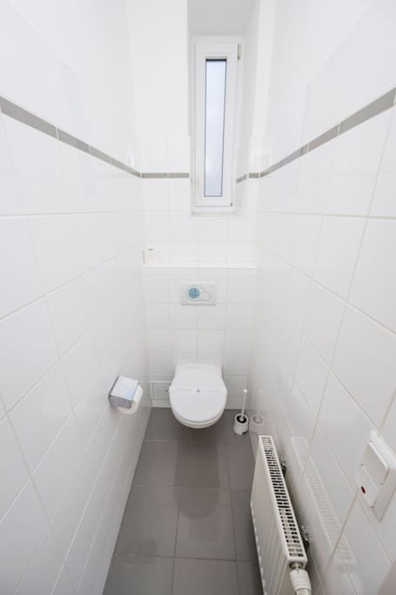 Une petite salle de bain étroite, avec des carreaux blancs, des toilettes, une fenêtre et un radiateur.