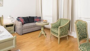A room with a bed, a grey sofa, a small round table with a bowl of fruit, a green patterned armchair, and a window with curtains. - Best of Hernals Top 30 (Vienna)