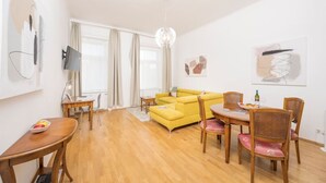 A modern living room with a yellow sofa, wooden dining table, and chairs. There is a TV mounted on the wall, a chandelier, and artwork on the walls. - Best of Hernals Top 34 (Vienna)