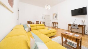 A living room with a yellow sofa, a wooden coffee table, a TV, and a dining area in the background. - Best of Hernals Top 34 (Vienna)