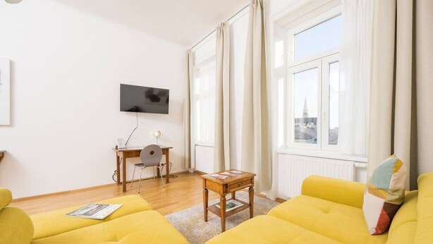 A bright living room with yellow sofas, a flat-screen TV, and a view of a cityscape through large windows. - Best of Hernals Top 34 (Vienna)