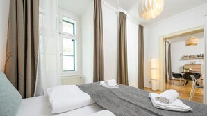 A hotel room with a bed, a desk, and a chair. There are curtains on the windows and a lamp on the desk.