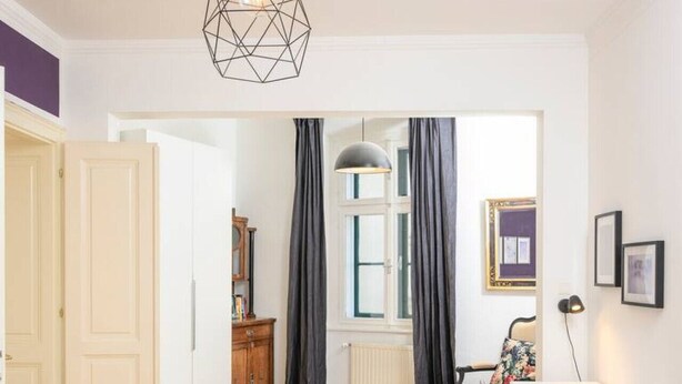 Une chambre moderne avec un lit, un bureau et une chaise. On y trouve une fenêtre avec des rideaux, un radiateur et une porte qui donne sur une autre pièce.