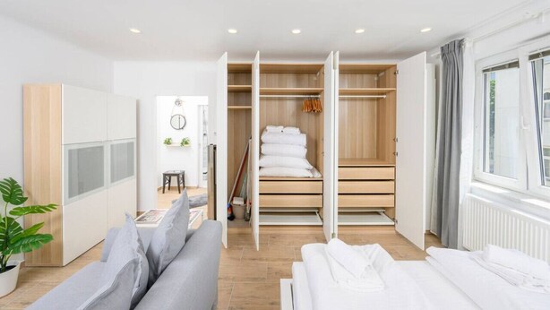 Une chambre d’hôtel moderne avec un canapé gris, un lit soigneusement fait, une armoire en bois et une vue sur l’extérieur grâce à une grande fenêtre.
