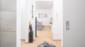 Un intérieur moderne avec un couloir menant à un espace de vie. On y trouve un porte-manteau avec quelques vêtements, un canapé gris et une petite table sur laquelle est posée une plante.