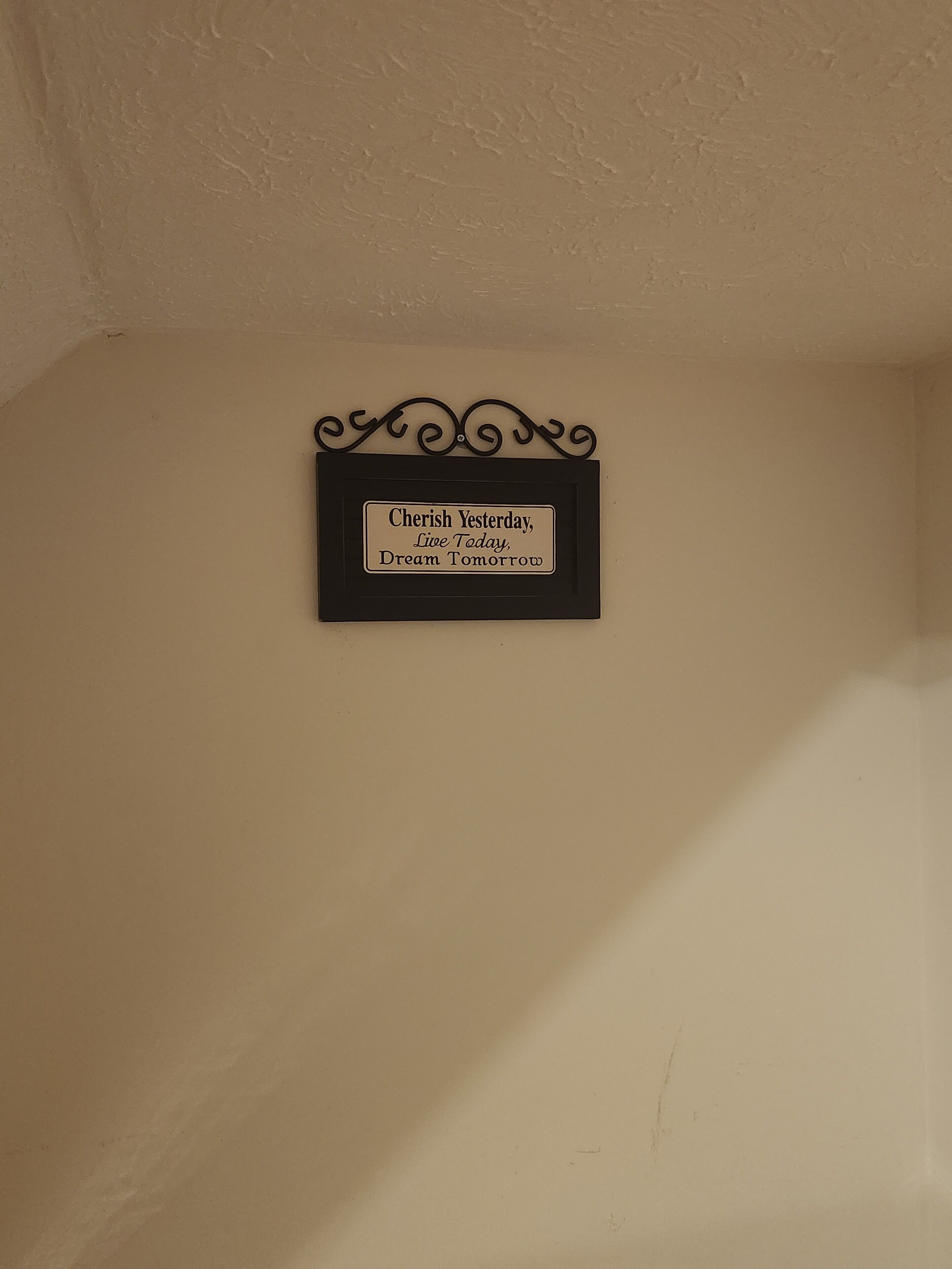 A wall-mounted sign with the text "Cherish Yesterday, Live Today, Dream Tomorrow" in a black frame with decorative top.
